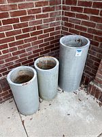 Three tall cylindrical ribbed metal planters with rust and dents, shown against a red brick exterior wall on a concrete floor, viewed from a slight angle.