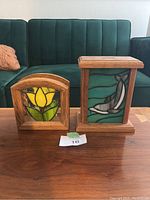 Photo showing two stained glass holders on a wooden table in front of a green couch. Left item is a napkin holder with yellow flower design, right item is a rectangular coin holder with bird design.