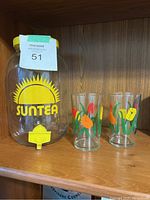 Photo showing glass beverage dispenser with yellow lid and spigot along with two floral glass tumblers on wooden shelf.