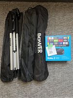 Three Bower tripods in carrying bags next to sealed Roku 2 XD box on grey textured surface.