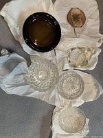 Five assorted ashtrays arranged on white cloth, 2 ceramic (one dark brown, one light brown) and 3 clear glass with various decorative patterns.