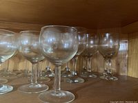 Twelve clear stem glasses of varied sizes arranged on wooden shelves, viewed from front showing rounded bowl shapes.