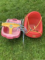 Two plastic baby swings on grass, one red with high back and one pink with yellow T-bar, both with ropes attached.