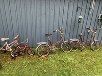 Four bikes shown including two brown folding bikes and two children's bikes (one red, one pink) on grass in front of a gray metal wall