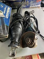 Photo showing two handheld power tools: a black Mastercraft angle grinder and a blue Mastercraft jigsaw. Both have power cords attached and some signs of use.