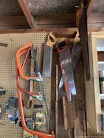 Photo shows four hand saws mounted on pegboard in a workshop. Two are orange framed bow saws and two are hand saws with wooden handles. The saws show some rust and wear.