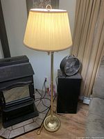 Tall brass floor lamp with beige pleated lampshade standing beside a small black stove and a speaker.