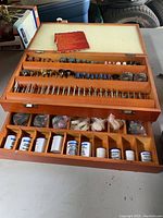 Wooden boxed set containing multiple rows of rotary tool bits including grinding stones, sanding drums, wire brushes, cutting discs, and polishing compounds in bottles and packets.