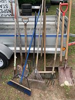 Photo of four wooden-handle shovels, one metal rake with a wooden handle, and one broom with blue plastic bristles and metal handle all leaning against a trailer.