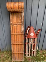 Photo of two wooden sleds leaning against a grey wooden wall. One sled has a natural wood finish and rope pull, the other has red metal cone and straps with wooden runners.