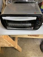 Compact silver and black toaster oven on a table, showing front glass door and control knobs.
