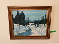 Framed winter landscape painting showing snowy ground, a flowing stream, pine trees, and a cabin or fire in the distance.
