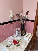 Bathroom counter with pink and black tile wall, displaying a tall black vase with faux calla lilies, soap dishes, small trinket boxes, white metal towel holder, small brown framed mirror, and white Kleenex tissue box cover.