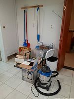 Wide view showing Mastervac vacuum cleaner on floor, several mops and brooms hanging on wall, metal shelving unit, garbage bin, and boxes with painting supplies and storage items