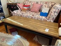 Full view of rectangular coffee table in front of floral couch with various boxes and cushions. Table top is laminate with wood grain pattern, base painted black.