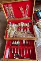 Photo shows wooden box lined with red velvet containing assorted gold colored Northcraft flatware mostly in plastic sleeves, and stainless steel Roberts and Son cutlery arranged neatly in compartments.