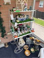 Front view of white metal vintage plant stand with three shelves, showing decorative hairpin legs and signs of paint chipping and rust. The shelves have green artificial grass patches and assorted decorative items, which are not included in the lot.