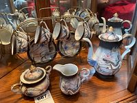 Full tea set displayed on wooden surface with reflective mirror background showing teapot, sugar bowl, creamer, cups hanging on metal stand, and saucers beneath.