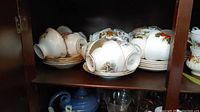 Multiple stacked porcelain teacups with matching saucers and floral gold-trim designs on a dark wooden shelf.