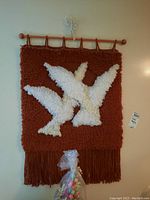 Front view of the handmade wall hanging rug on wooden hanger showing white bird design and fringes with bag of colored items in foreground.