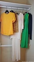 Five clothing items hanging on a rod: yellow shirt, gold sequin shirt, white dress, green dress, black cardigan.