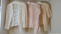 Three vintage lace jackets hanging on hangers; visible delicate lace patterns; colors cream and light pink.