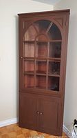 Full view of the closed corner china cabinet showing wooden frame, glass door with grid pattern, and two knobs on bottom doors.