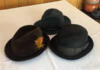 Three felt hats laid side by side on a white surface: a dark gray fedora with braided band, a brown fedora with a feather in band, and a black bowler hat with wide ribbon.
