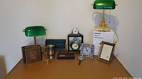 Full view of lot on desk, showing 3 green Tensor bankers desk lamps, 2 clocks, picture frames, magnifying glass, mug and other small items