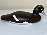 Side view of Harold Levy wooden duck decoy showing dark brown body, white wing patch, and blue bill.