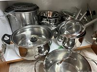 Overall view of stainless steel cookware including loop-handle pots with lids, saucepans, mixing bowls, stockpot, and grilling mats