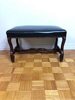 Front view of leather bench showing cushioned seat, nailhead trim, and carved wooden legs with small casters.