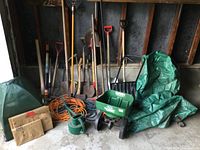 Photo showing various long-handled garden tools including rakes, hoes, shovels; green seed spreader; watering can; orange extension cords; green tarps and cardboard mailers arranged against wall