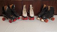 Three pairs of vintage roller skates displayed on a white surface against a wooden background: two pairs of black Bauer skates with red wheels and one pair of white roller skates with red wheels.