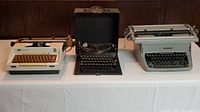 Three mechanical typewriters arranged on table: a grey Model 5, black Remington Portable Model 5 in case, and Office Electric "Coronamatic" typewriter