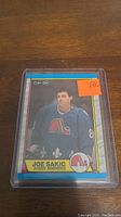 Joe Sakic rookie hockey card in O-Pee-Chee brand featuring Quebec Nordiques jersey and logo, in protective casing to prevent damage.