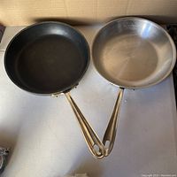 Two frying pans side by side on a white surface: one black non-stick, one stainless steel with metal handles.