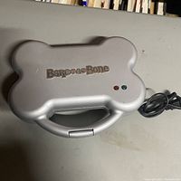 Closed view of silver dog treat maker shaped like a bone with brand logo and indicator lights on top, power cord visible.