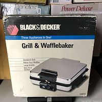 Front view of packaging showing product name 'Grill & Wafflebaker' with features: Sandwich Grill, Bakes Four Waffles, Large Griddle, Non-Stick Surface