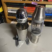 Hamilton Beach electric mixer with attached stainless steel cup next to Belvedere 007 branded cocktail shaker on table with bookshelf background