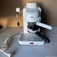Front view of white Krups espresso machine powered on with attached portafilter and glass carafe underneath, stainless steel drip tray, and metal steam wand with black tip.