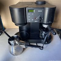Front view of the black Krups espresso machine with control buttons and digital screen, accompanied by the frothing pitcher and coffee scoop in front.