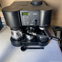 Front view of black Krups espresso coffee machine with portafilter attached, digital controls, and accessories including milk frothing pitcher, scoop, and tamper placed in front.
