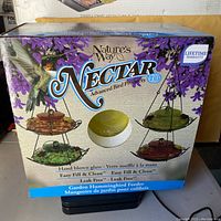 Front view of Nature's Way Nectar Series Garden Hummingbird Feeder box, showcasing the hand blown glass feeder in red, yellow, green, and purple options with lifetime warranty noted.