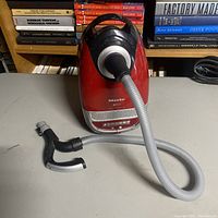 Front angle showing red vacuum body with attached gray hose and black handle, books in background.