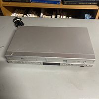 Top view of silver Samsung DVD VCR combo unit showing front control buttons and display panel, power cord coiled behind unit.