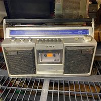 Front view of Sony portable boombox showing dual speakers, cassette deck, control knobs, and blue AM/FM dial.