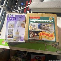 Photo showing Wilton Simple Success Cookie Press in its box and Hamilton Beach Deluxe SuperShooter in original packaging side by side on a table.