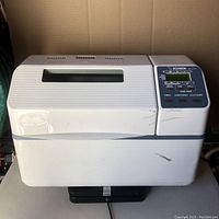 Front view of the white Zojirushi bread maker with visible control panel and top viewing window, showing some surface scratches and marks.