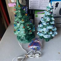 Photo showing both ceramic Christmas trees, one with snow effect, with a bag of extra plastic light bulbs and an electrical cord on table.